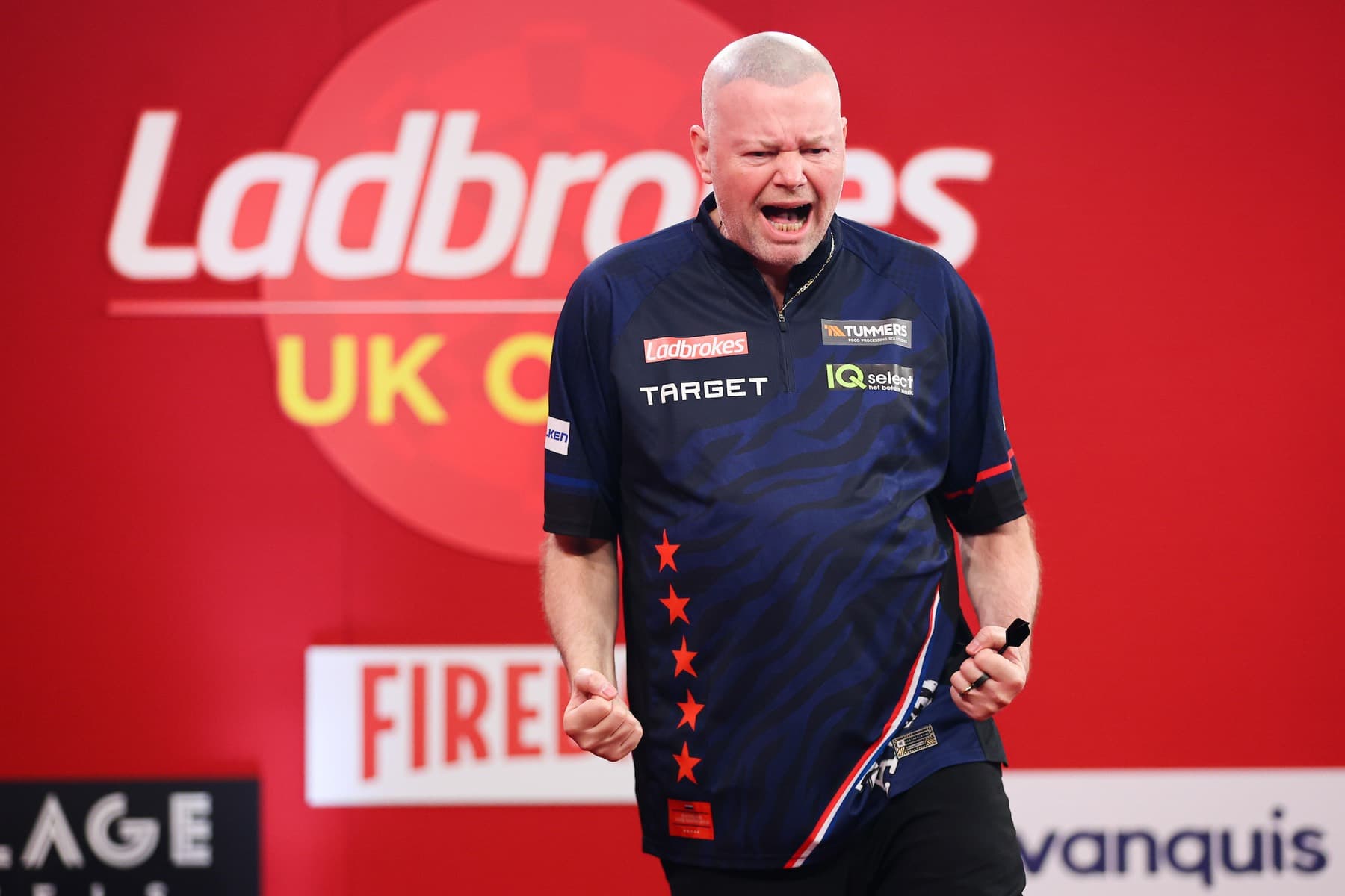 Raymond Van Barneveld celebrating on stage with clenched fists in front of a red backdrop