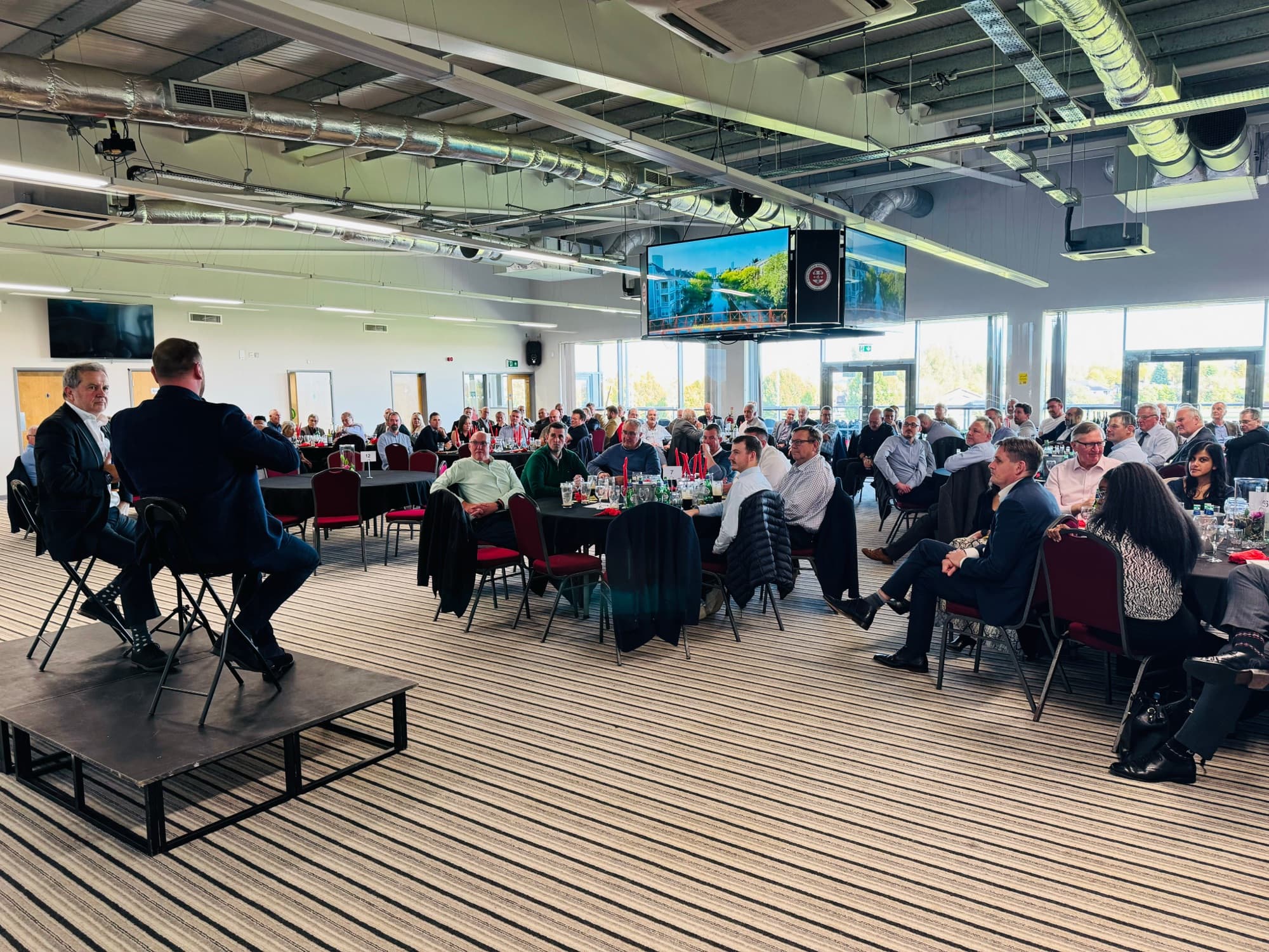 The Reddings Suite set with round tables for an event, with a stage and full-length windows over the pitch