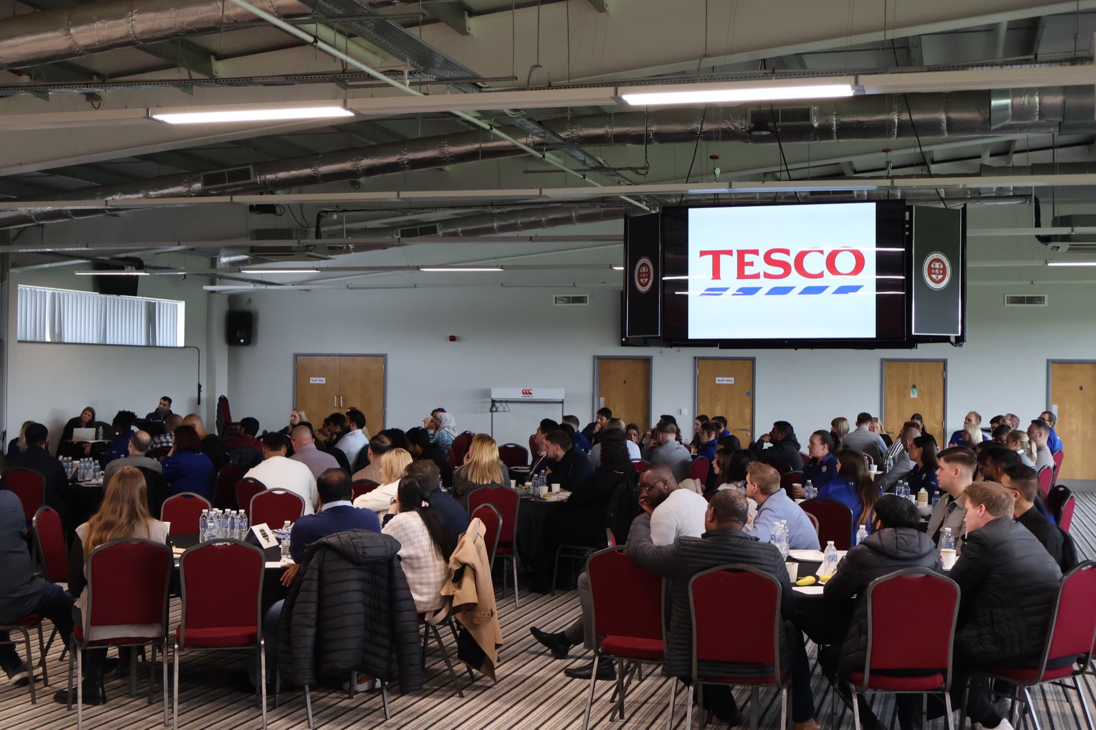 A packed corporate conference for Tesco in The Reddings Suite, delegates at round tables with the Tesco logo on the big screen